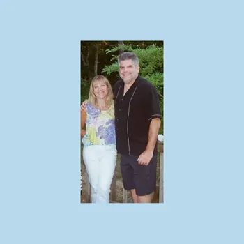Women standing on the left and man of the right both widely smiling with shrubbery in the background.