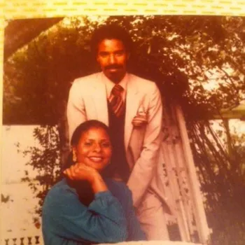 Older colored photo of man leaning back on a trellis in a garden and a women squatting in front of him both smile for photo.