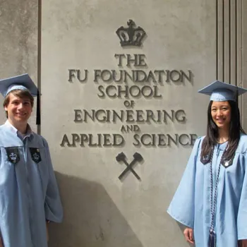 Male standing left and women standing right both in alumni robes between the Fu Foundation School statue.