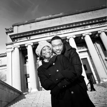 A young couple playfully smiling and hugging with the man hugging the backside of a women while they face the camera -- the backdrop is low library.