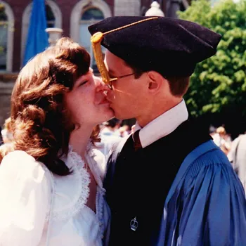 A women on the left dressed in white flowery dress and a man on the right wearing alum robes and a tassel kissing at graduation.