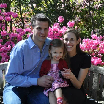 Male and female couple and their female toddler, sitting in a park with flowers in the background.