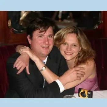 A man and woman sit at a table, hugging, while a camera rests between them.