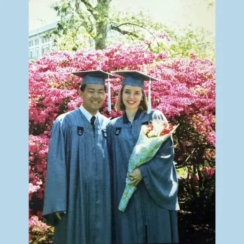 Man and woman in Columbia cap and gown, standing in front of a flowering bush. The girl holds a bunch of wrapped flowers.