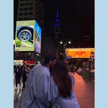 Man and woman in their Columbia Blue graduation robes, walking through Times Square with the Empire State Building in the background, lit in Columbia blue.