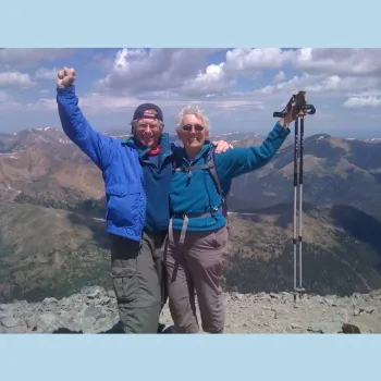 Two people stand triumphantly on a mountain peak, arms raised in celebration against a clear blue sky.