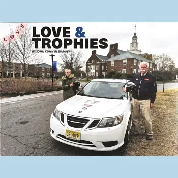 Man woman standing by a white sports car, in front of a house in a rural setting. Print on the image reads "Love & Trophies."