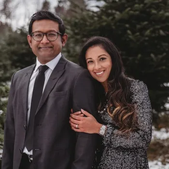 Man and woman standing close together outside by pine trees.