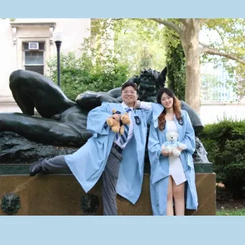 Two individuals in robes in Columbia commencement regalia pose for a photo in front of a statue, smiling and standing side by side.