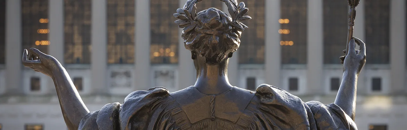 Statue of ALMA with raised arms in front of Butler Library at dusk.