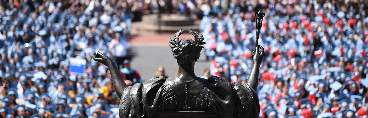 A bronze statue with outstretched arms faces a large crowd of people in blue graduation gowns and caps at an outdoor ceremony.
