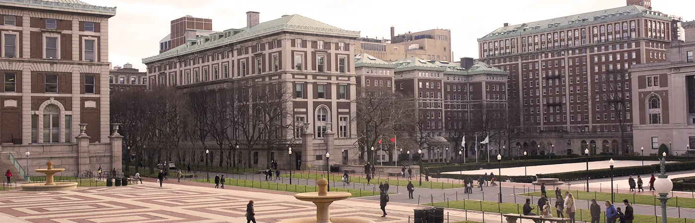 Columbia University campus with historic buildings, a central lawn, and people walking across the plaza.