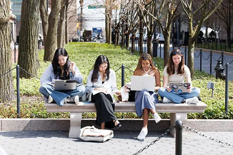Four students sitting on a bench using laptops along a tree-lined campus walkway