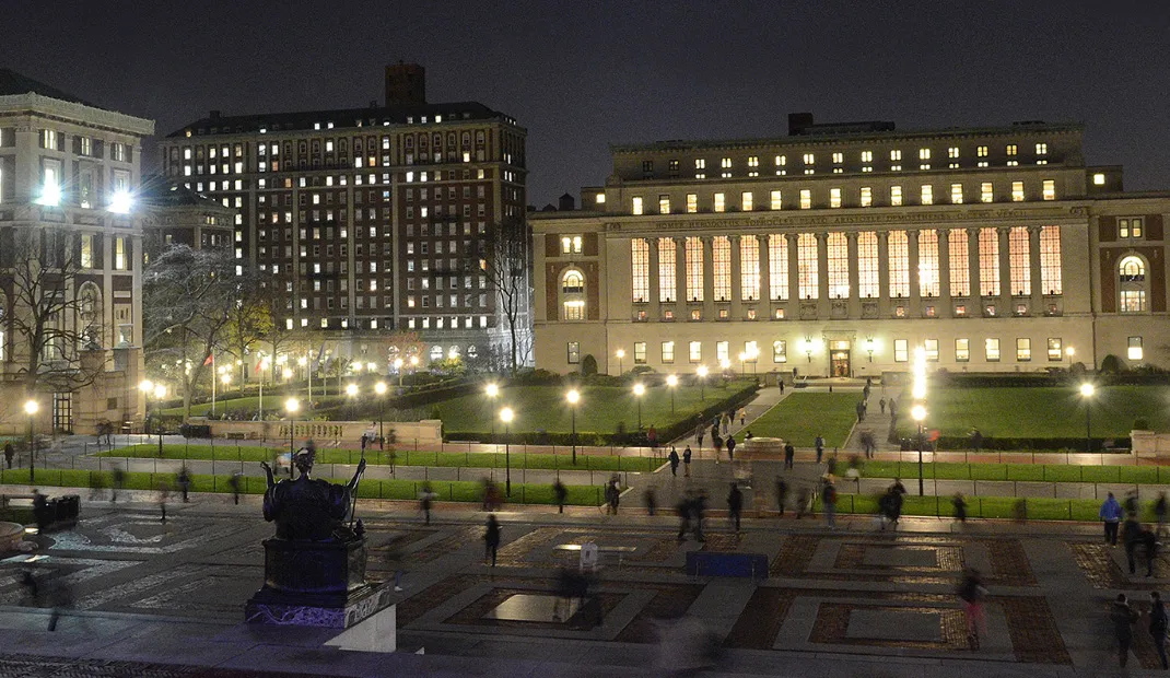 Butler Library at Columbia University illuminated at night, with people walking across the campus plaza.