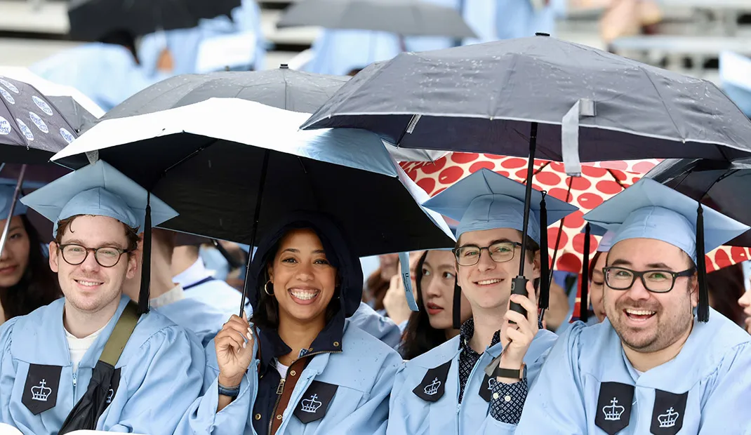 Graduates in caps and gowns holding umbrellas in the rain, smiling during the Columbia University Commencement 2025 ceremony.